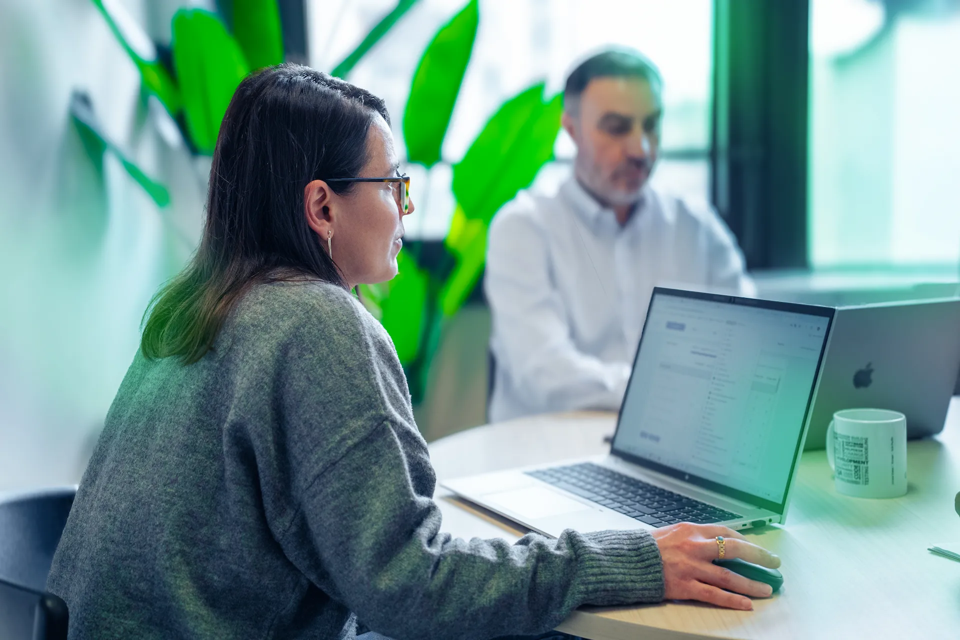 Professionista di Winet al laptop, concentrata su soluzioni software per Thales, mentre un collega è seduto di fronte in un ufficio moderno con piante verdi.